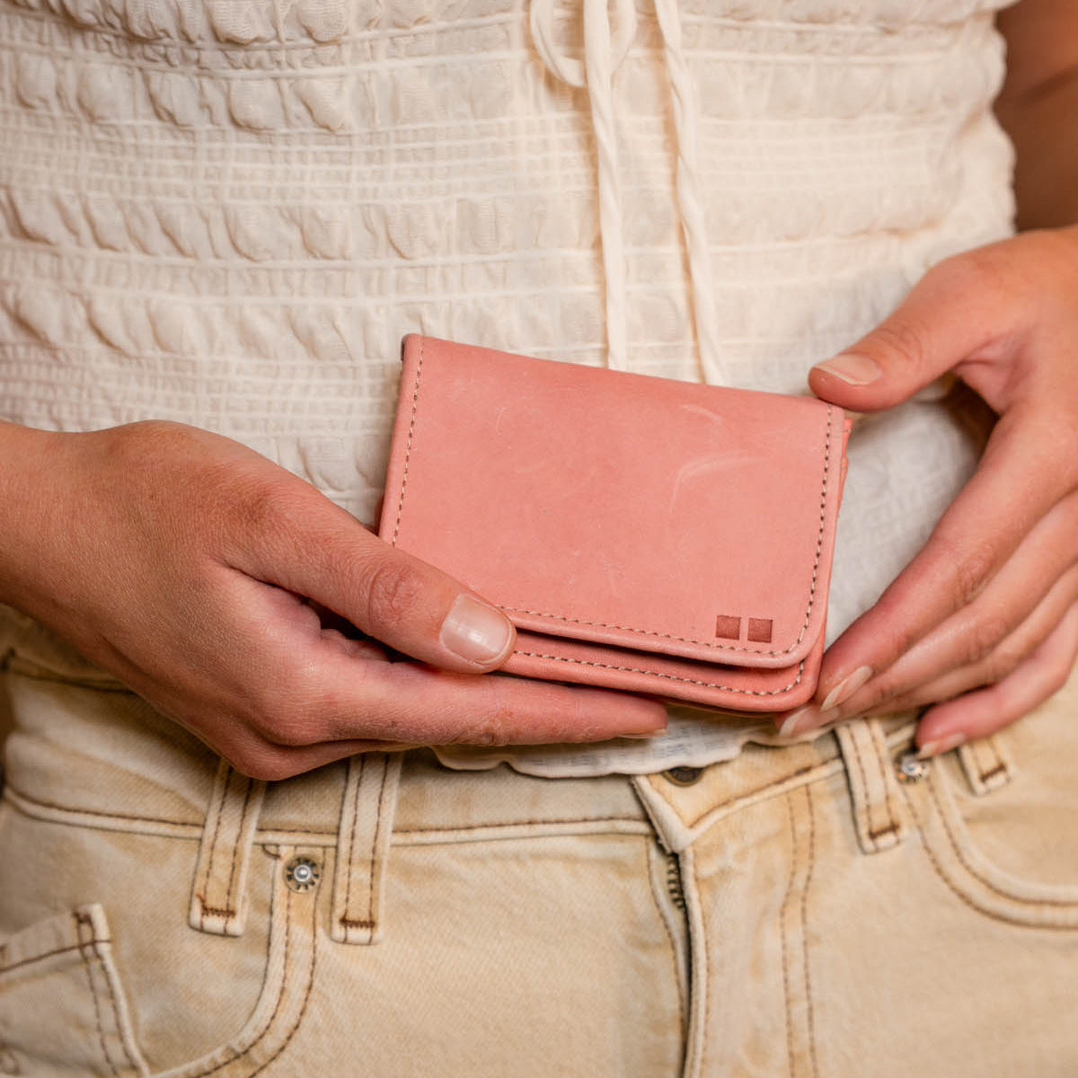 A person holds the Bed Stu Jeor wallet in light pink with both hands, showcasing its multiple credit card slots, while dressed in a white textured top and beige pants.