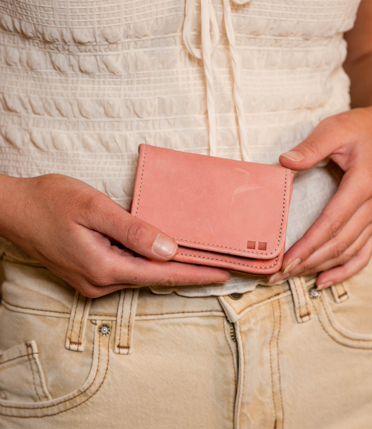 A person holds the Bed Stu Jeor wallet in light pink with both hands, showcasing its multiple credit card slots, while dressed in a white textured top and beige pants.
