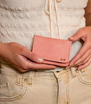 A person holds the Bed Stu Jeor wallet in light pink with both hands, showcasing its multiple credit card slots, while dressed in a white textured top and beige pants.