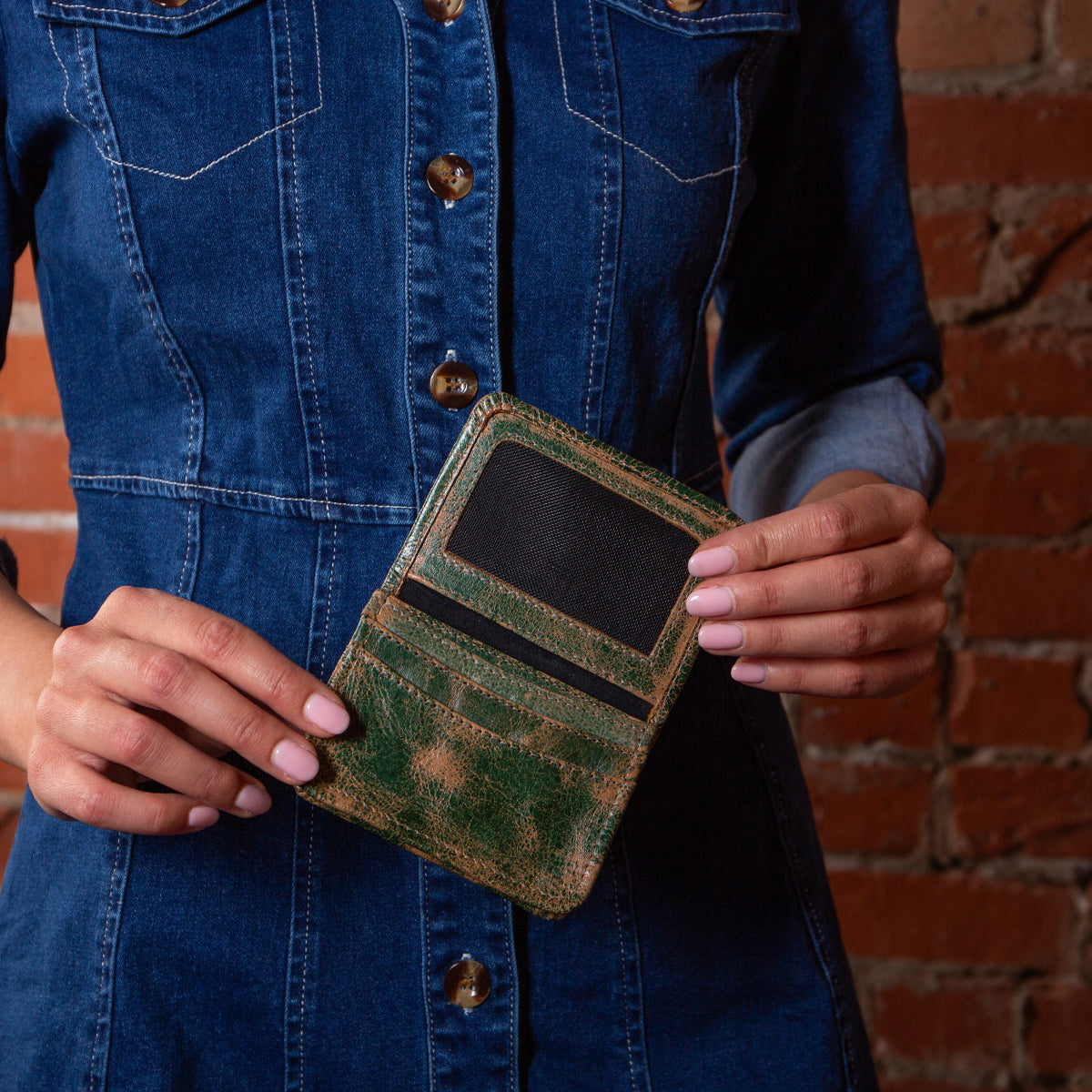 A person in a blue denim dress holds the Bed Stu Jeor, an open rustic leather card holder with visible card slots, against a brick wall background.