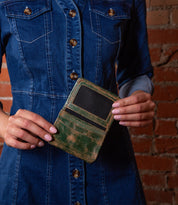 A person in a blue denim dress holds the Bed Stu Jeor, an open rustic leather card holder with visible card slots, against a brick wall background.