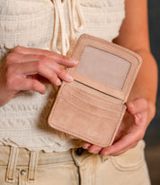 A person holds the Bed Stu Jeor, a compact wallet with credit card slots and a transparent ID window, while wearing a white textured top and beige pants.