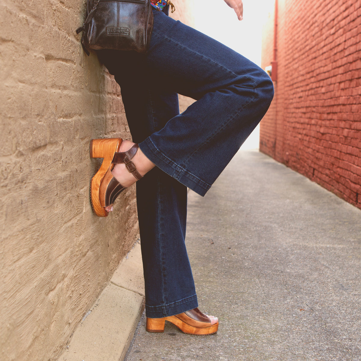 A person wearing Bed Stu's Jetsetter flared jeans and brown platform shoes with patchwork finishes leaning against a brick wall.