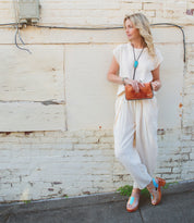 A woman in a white outfit with a turquoise necklace stands by a textured wall with patchwork finishes, holding a Bed Stu Jetsetter clutch.