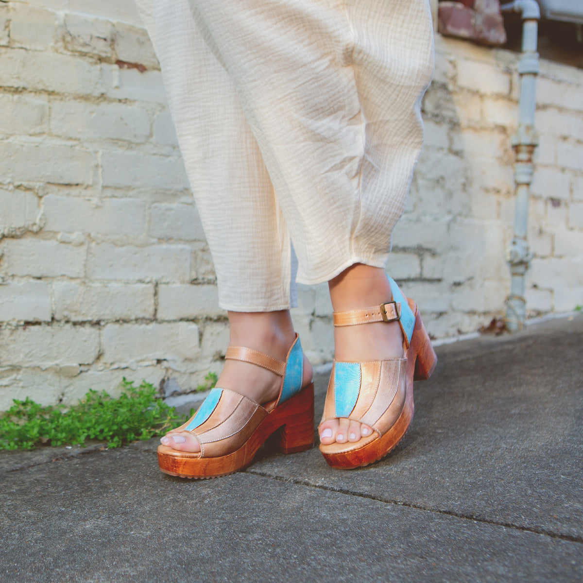 A person standing in Bed Stu's Jetsetter leather heels with patchwork finishes on a pavement next to a wall.