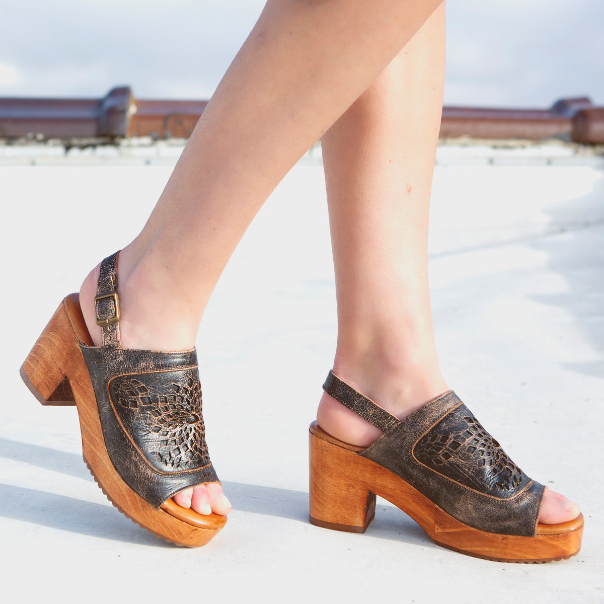 A woman wearing a pair of Bed Stu Jinkie wooden sandals on a roof. Decorating her feet with open-toe leather sandals, she confidently walks across the rooftop, the heeled wooden platform providing a stylish lift.