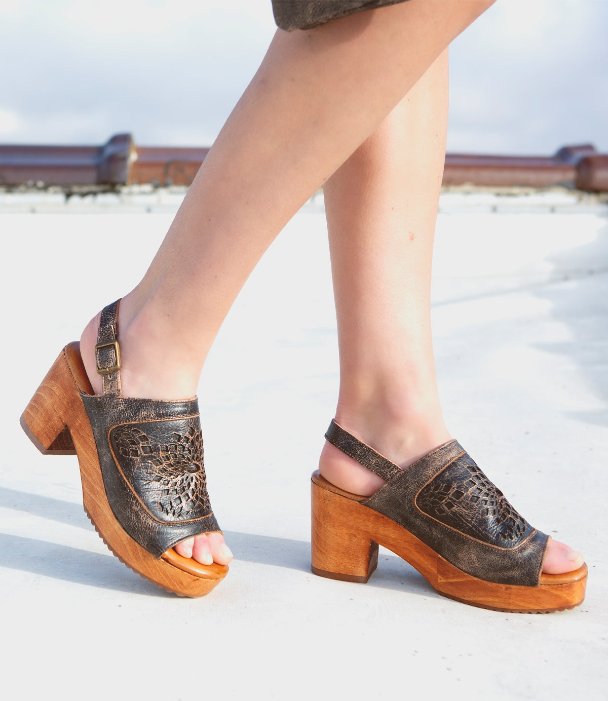 A woman wearing a pair of Bed Stu Jinkie wooden sandals on a roof. Decorating her feet with open-toe leather sandals, she confidently walks across the rooftop, the heeled wooden platform providing a stylish lift.