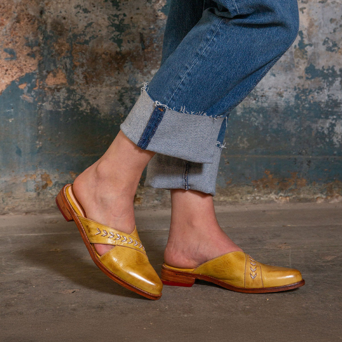Wearing Bed Stu’s Jockey—yellow slip-on leather shoes with arrow inlays—a person in rolled-up blue jeans stands on a concrete floor, their standout style enhanced by the weathered wall behind them.