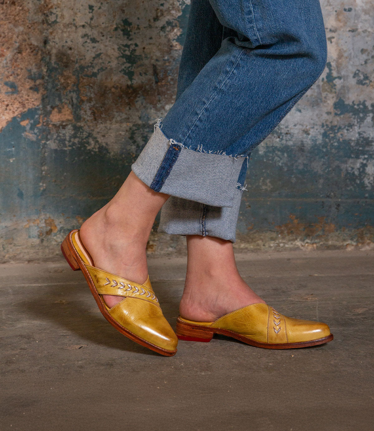 Wearing Bed Stu’s Jockey—yellow slip-on leather shoes with arrow inlays—a person in rolled-up blue jeans stands on a concrete floor, their standout style enhanced by the weathered wall behind them.