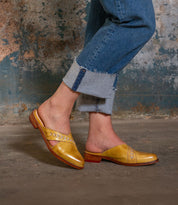 Wearing Bed Stu’s Jockey—yellow slip-on leather shoes with arrow inlays—a person in rolled-up blue jeans stands on a concrete floor, their standout style enhanced by the weathered wall behind them.