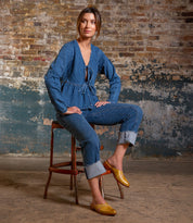 A woman sits on a chair in front of a textured brick wall, wearing the Bed Stu Jockey yellow slip-on shoes for standout style, paired with a blue long-sleeve blouse and cuffed jeans.