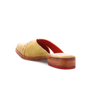 The Bed Stu Jockey is a tan leather slip-on mule with a sleek design, wooden heel, and red sole, shown from the back on a white background.