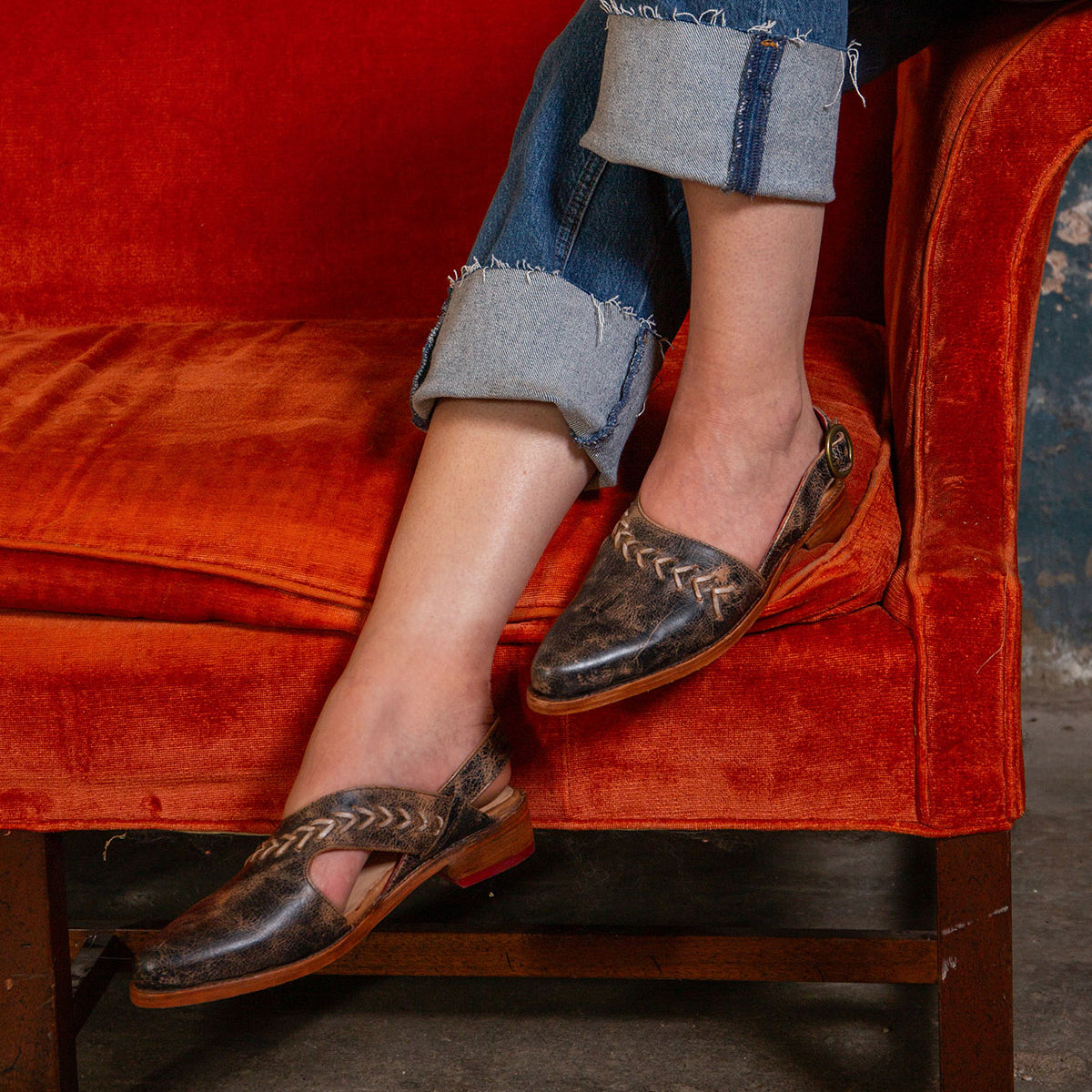 A person wearing Bed Stu Join brown leather sling back flats with an adjustable buckle sits cross-legged on an orange velvet sofa, paired with distressed jeans.