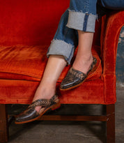 A person wearing Bed Stu Join brown leather sling back flats with an adjustable buckle sits cross-legged on an orange velvet sofa, paired with distressed jeans.