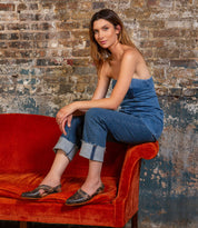 A woman wears the Bed Stu Join strapless denim jumpsuit with adjustable buckle, sitting sideways on a red velvet couch against a weathered brick wall and looking at the camera.