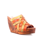The Bed Stu Josie II is a wooden wedge sandal featuring hand-woven orange and yellow leather straps with metal studs, shown on a white background.