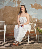 A woman in a white dress and Bed Stu Josie II wooden wedge sandals with metal studs sits on a white metal chair against a textured wall, with a vase of flowers on the floor nearby.
