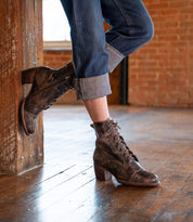 A person in cuffed blue jeans and Bed Stu Judgement boots with a lace-up design and block heel leans against a wooden post on a wooden floor with exposed brick walls.