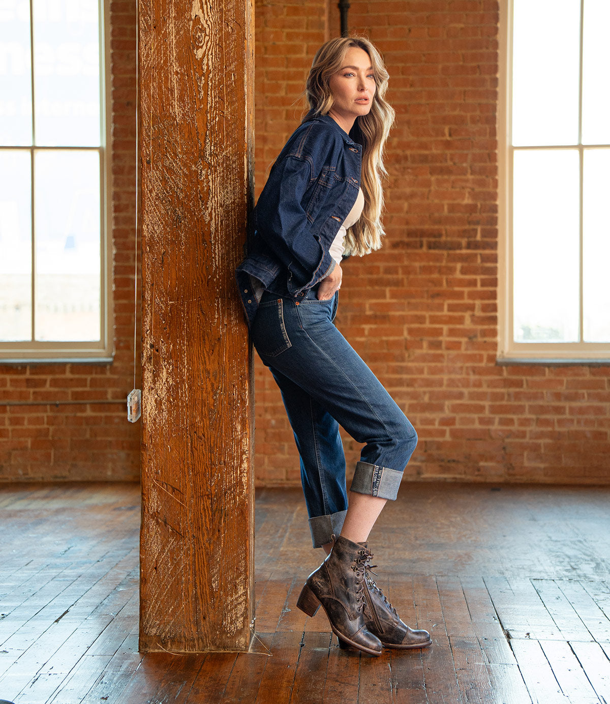 A woman with long hair in a denim jacket and jeans leans against a wooden pillar, showcasing Bed Stu Judgement 8-eye lace-up boots with a block heel in a room featuring brick walls and large windows.