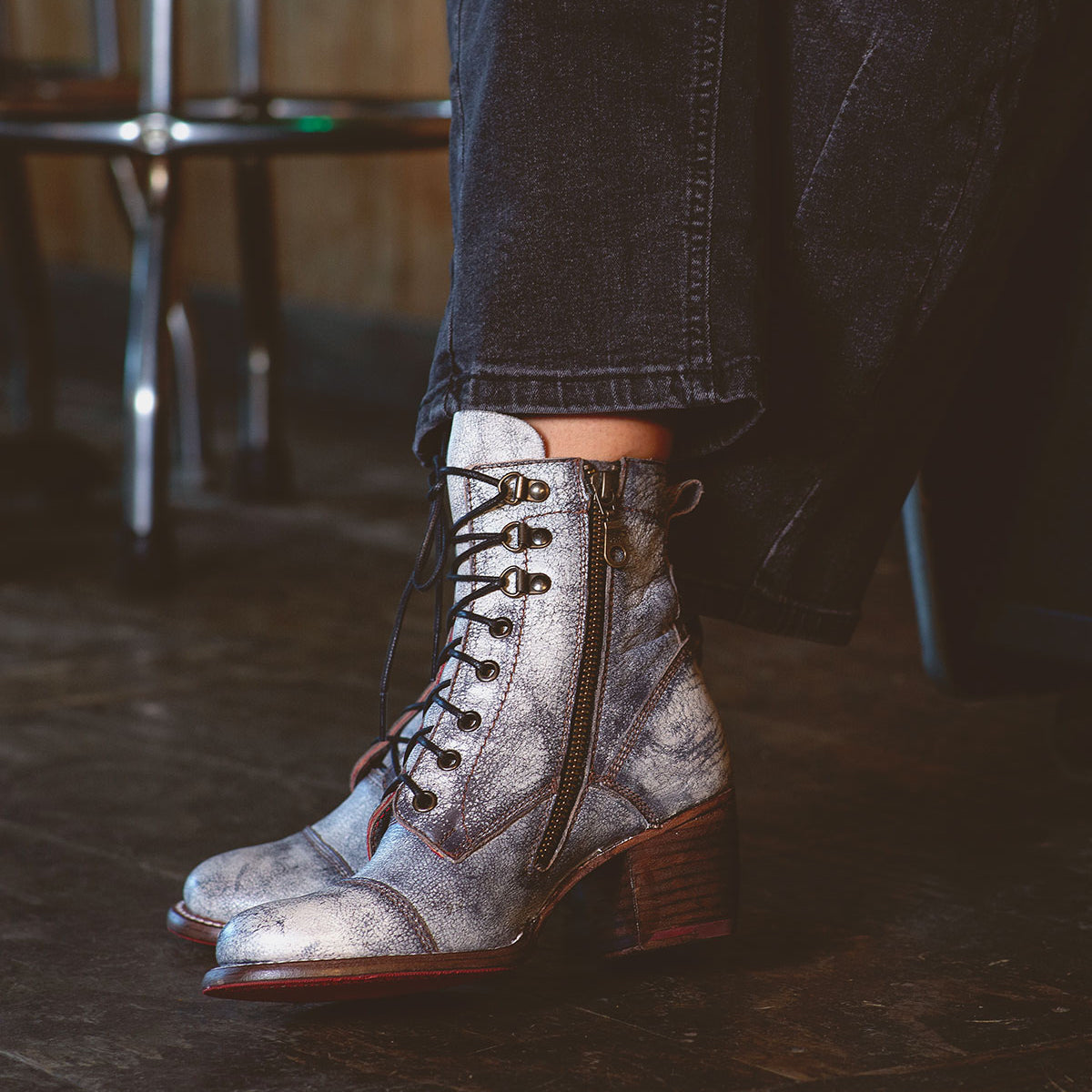 A person is standing on a wooden floor, sporting Bed Stu's Judgement boots, which feature metallic finishes, a zipper, and cushioned insoles for added comfort, paired chicly with dark pants.