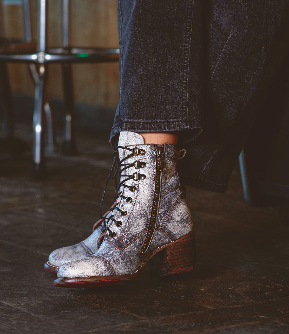 A person is standing on a wooden floor, sporting Bed Stu's Judgement boots, which feature metallic finishes, a zipper, and cushioned insoles for added comfort, paired chicly with dark pants.