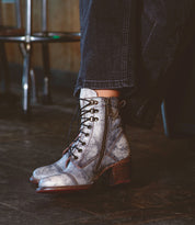 A person is standing on a wooden floor, sporting Bed Stu's Judgement boots, which feature metallic finishes, a zipper, and cushioned insoles for added comfort, paired chicly with dark pants.