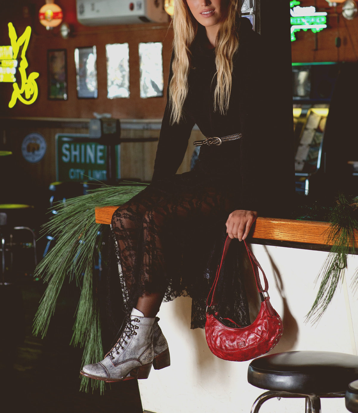 A person in a black lace dress sits on a wooden bar counter, holding a red handbag. They wear the Judgement 8-eye lace-up boots by Bed Stu, featuring cushioned insoles. Pine branches decorate the area, while neon signs glow in the background.