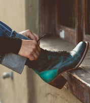 A person laces up a teal and black Bed Stu Judgement boot, featuring an 8-eye lace-up design, while seated next to a wooden windowsill.
