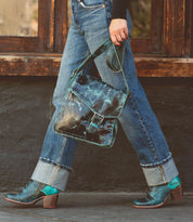 A person wearing cuffed jeans and ankle boots with a cushioned insole carries Bed Stu's Judgement, a worn blue leather satchel, over their shoulder while walking on a sidewalk.