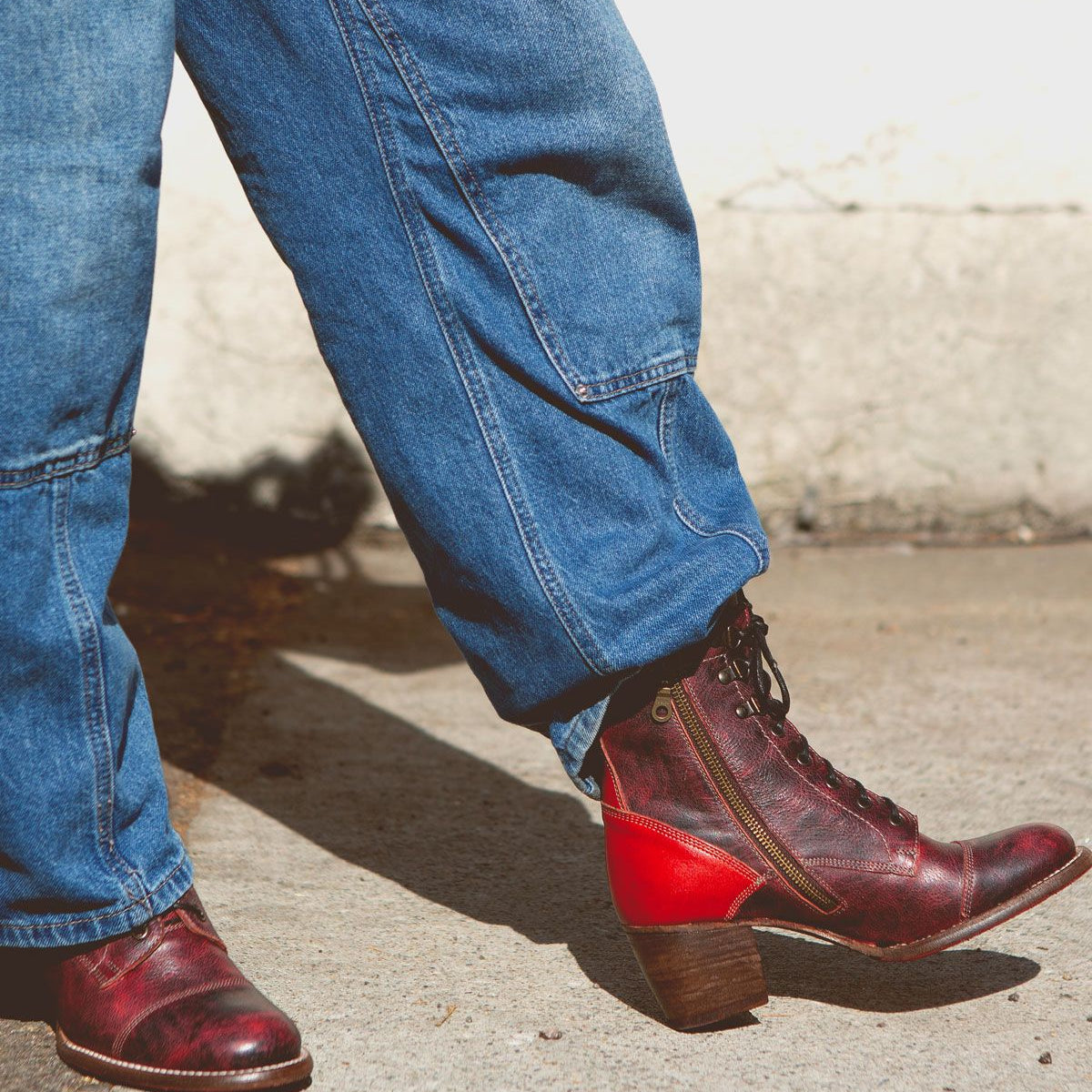 A person wearing blue jeans and brown Bed Stu Judgement boots with an 8-eye lace-up design is standing on a concrete surface in natural light.