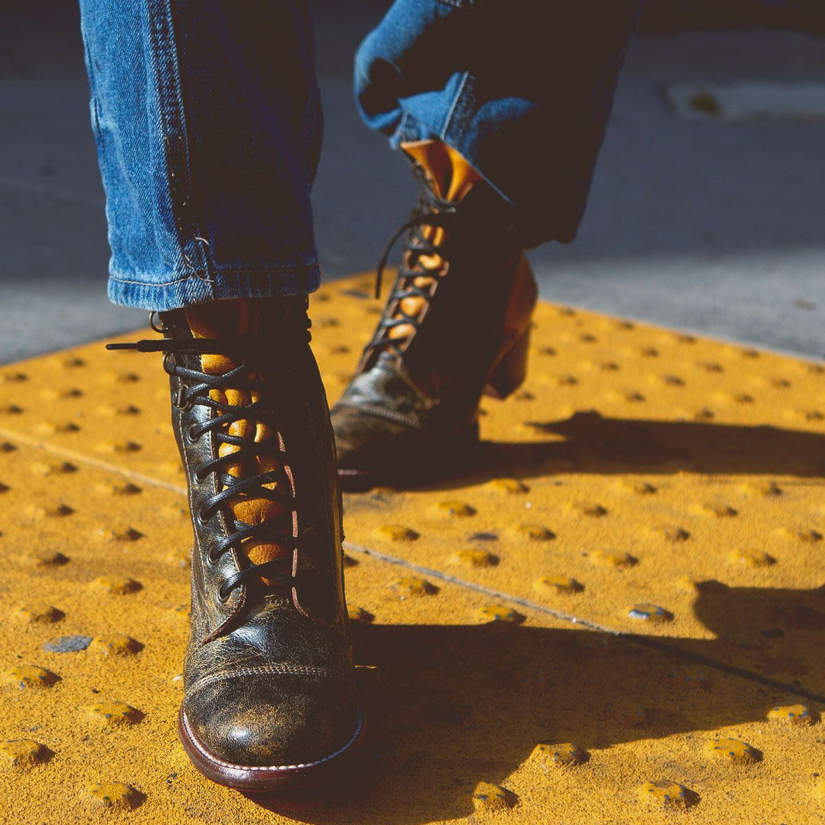 Close-up photo of a person wearing Bed Stu Judgement boots with an 8-eye lace-up design and blue jeans, standing on a yellow textured surface.