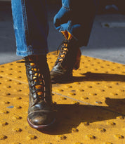 Close-up photo of a person wearing Bed Stu Judgement boots with an 8-eye lace-up design and blue jeans, standing on a yellow textured surface.