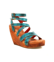 The Bed Stu Juliana II is a teal platform sandal with a strappy upper, ankle buckle, and sleek wooden wedge heel, shown on a white background.