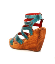 The Bed Stu Juliana II is a turquoise platform sandal with a strappy upper, wooden wedge sole, and metal rivet accents, shown in a side view.
