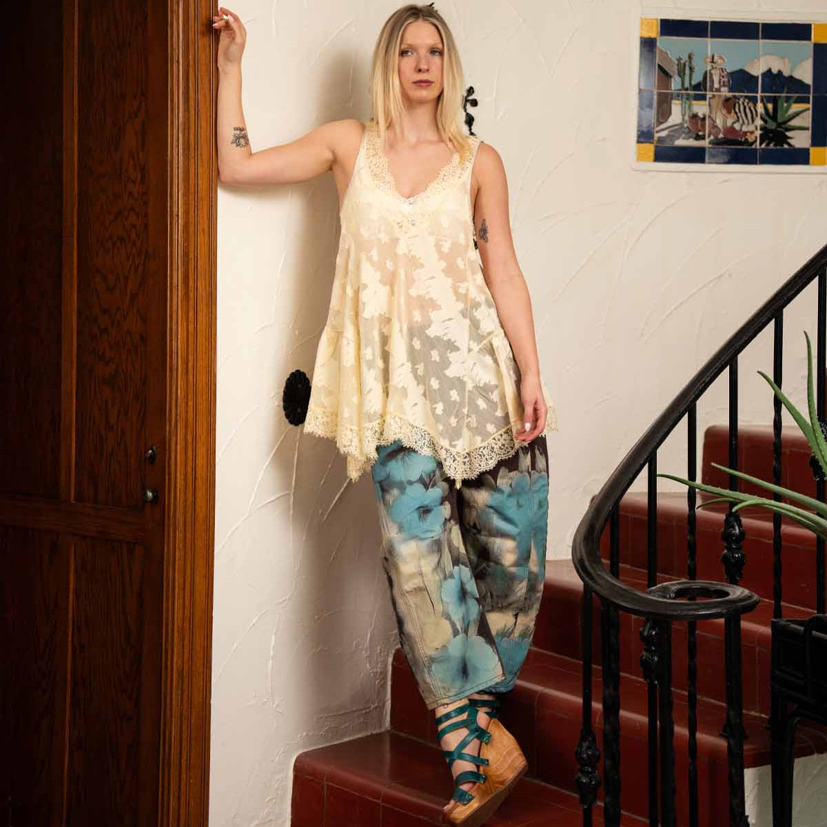 A woman in a cream lace top and blue patterned pants stands on red indoor stairs beside a white wall and wooden door, wearing Bed Stu's Juliana II platform sandals with a wooden wedge and strappy upper.
