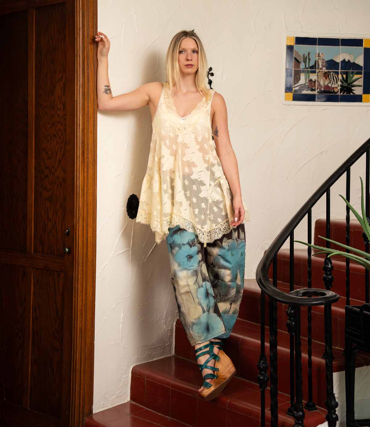 A woman in a cream lace top and blue patterned pants stands on red indoor stairs beside a white wall and wooden door, wearing Bed Stu's Juliana II platform sandals with a wooden wedge and strappy upper.