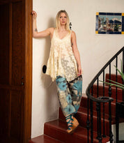 A woman in a cream lace top and blue patterned pants stands on red indoor stairs beside a white wall and wooden door, wearing Bed Stu's Juliana II platform sandals with a wooden wedge and strappy upper.