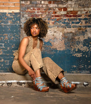 A woman with curly hair sits on a concrete ledge against a weathered brick wall, wearing tan overalls and blue Bed Stu Juliana II platform sandals with a wooden heel.