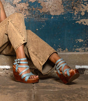 A person wearing tan jeans and Bed Stu Juliana II light blue strappy wedge sandals with cushioned soles and adjustable buckles sits against a weathered blue wall.