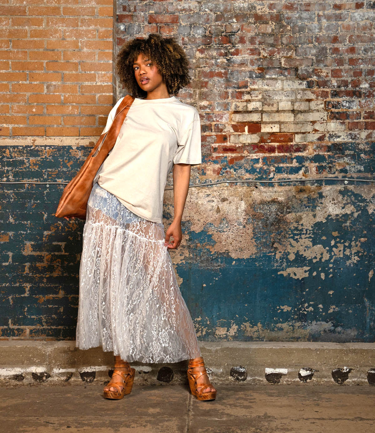 A person stands against a worn brick wall, wearing a beige t-shirt, sheer lace skirt, carrying a large brown bag, and wearing Bed Stu Juliana II brown sandals with wooden heels.