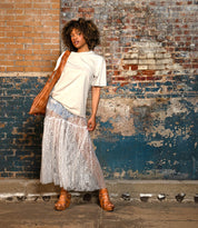 A person stands against a worn brick wall, wearing a beige t-shirt, sheer lace skirt, carrying a large brown bag, and wearing Bed Stu Juliana II brown sandals with wooden heels.