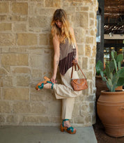 A woman stands by a stone wall, adjusting her Bed Stu Kalah blue platform sandal with an adjustable ankle strap. She pairs beige pants with a patterned top and brown purse, near large pots of cactus plants.