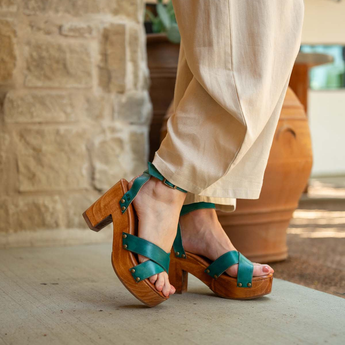 A person wears the Bed Stu Kalah teal platform sandals, featuring a sustainably sourced wooden heel, paired with beige pants, standing on concrete near a stone wall and large terracotta pots.