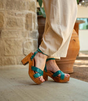 A person wears the Bed Stu Kalah teal platform sandals, featuring a sustainably sourced wooden heel, paired with beige pants, standing on concrete near a stone wall and large terracotta pots.
