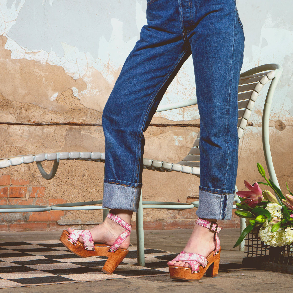 In cuffed jeans on a checkered floor, someone showcases Bed Stu's Kalah pink floral platform heels. A metal chair and a flower bouquet enhance the scene, reflecting eco-consciousness with sustainably sourced elements against a textured wall.