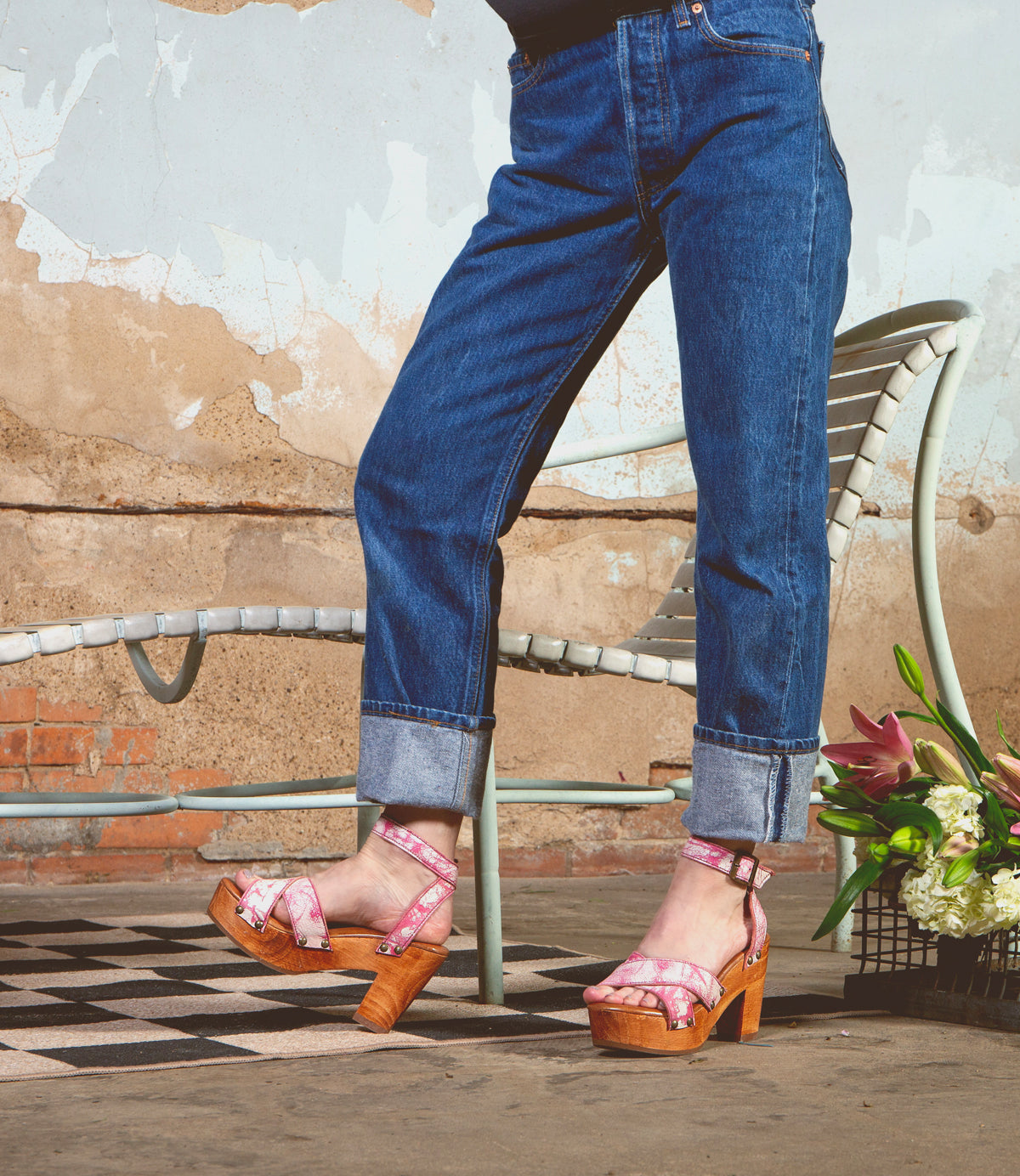 In cuffed jeans on a checkered floor, someone showcases Bed Stu's Kalah pink floral platform heels. A metal chair and a flower bouquet enhance the scene, reflecting eco-consciousness with sustainably sourced elements against a textured wall.