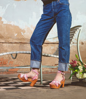 In cuffed jeans on a checkered floor, someone showcases Bed Stu's Kalah pink floral platform heels. A metal chair and a flower bouquet enhance the scene, reflecting eco-consciousness with sustainably sourced elements against a textured wall.
