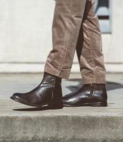 A person strolls on a concrete surface, donning a pair of Kaldi brown leather ankle boots by Bed Stu with a classic silhouette and beige pants.