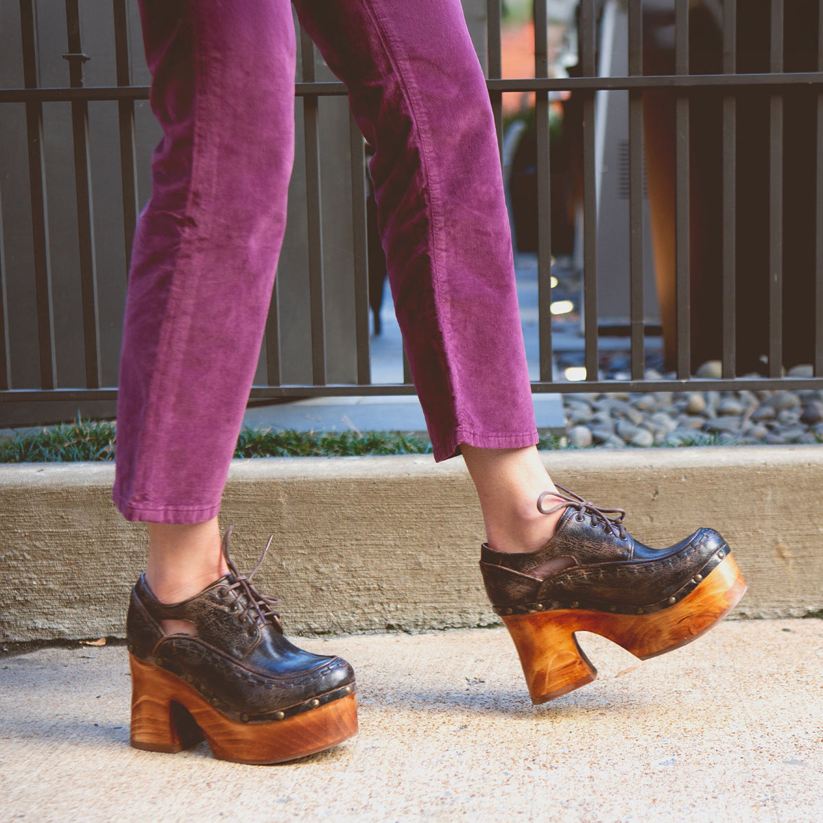 A person wearing Bed Stu's Karaoke purple pants and chunky wooden platform shoes, reminiscent of the 70s era, strides confidently down a concrete path.
