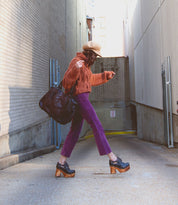 A person in a brown hat, orange sweater, and purple pants strolls through an alley carrying a large bag, sporting the *Karaoke* chunky wooden platforms from Bed Stu, reminiscent of the 70s era.